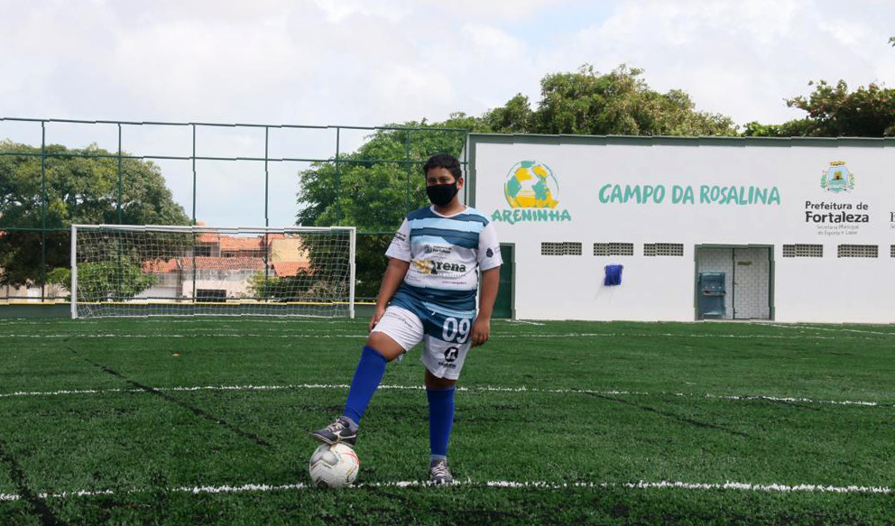 garoto em pé posando para a foto com a bola no pé no gramado da Areninha